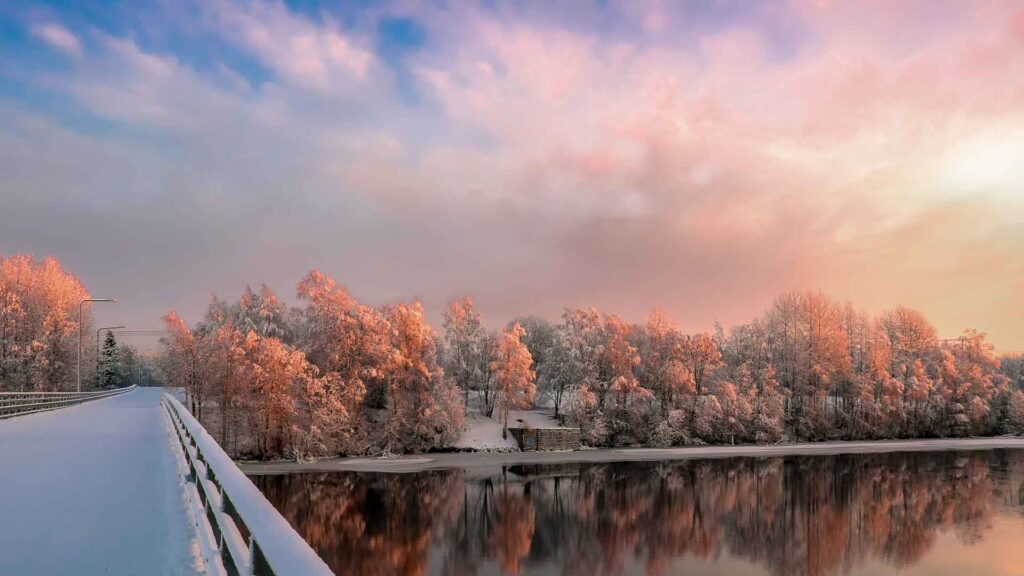 Snötäckta träd i orange sken speglas i vattnet där bron från Östteg korsa till Ön. En hälsning från  Ekmans Handelsträdgård.